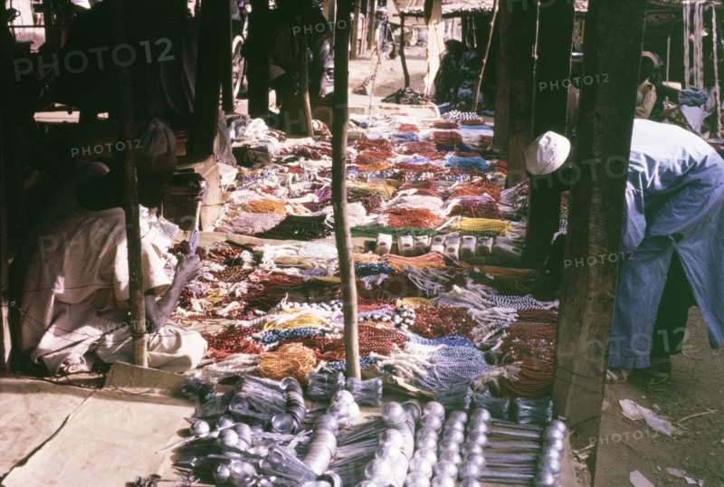 Beaded jewellery on a market stall. A customer bends down to inspect a huge selection of colourful beaded jewellery on a market stall. Kano, Nigeria, October 1963. Kano, Kano, Nigeria, Western Africa, Africa.