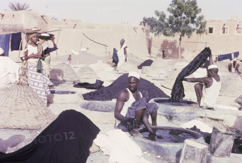Indigo dyeing at Kano. Female hawkers circulate amongst textile workers who sit outdoors, dipping lengths of fabric into wells of indigo dye. Several unused wells are covered with basketwork lids as protection against the sun. Kano, Nigeria, 1963. Kano, Kano, Nigeria, Western Africa, Africa.