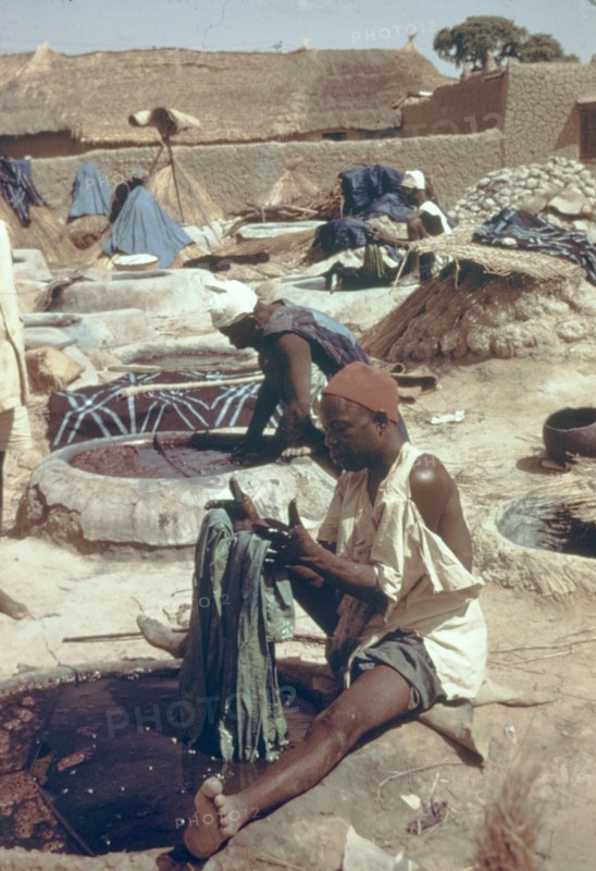 Hausa man dyeing fabric. A Hausa man sits beside a well of liquid dye, carefully dipping a section of fabric into the substance at an outdoor treatment workshop. Nigeria, circa 1975. Nigeria, Western Africa, Africa.
