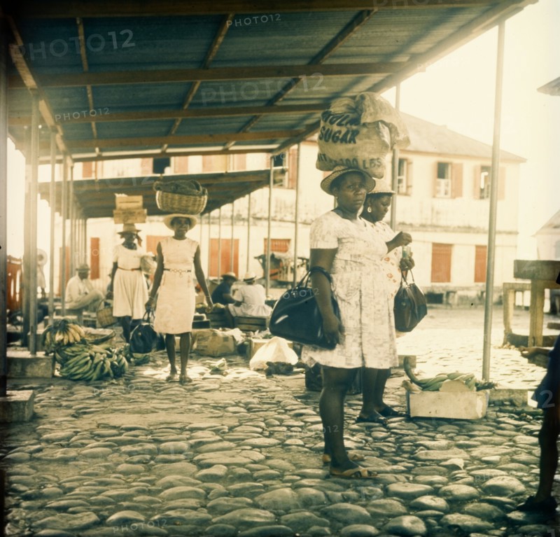 Dominican women at a street market. Dominican women at a street market carry their shopping in sacks, baskets and crates balanced on their heads. Dominica, circa 1975. Dominica, Caribbean, North America .