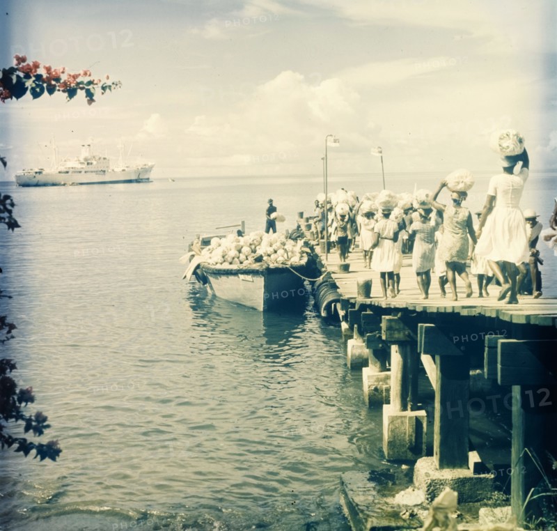 Maritime trade, Dominica. A group of women walk along a wooden pier carrying sacks on their heads, which they load into a motor boat waiting in harbour. A luxury liner is moored offshore, possibly waiting to receive the supplies. Dominica, circa 1975. Dominica, Caribbean, North America .
