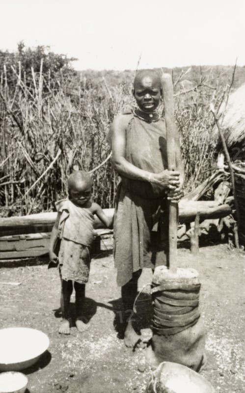 Preparing food outdoors, Kenya. A shy young child clings to his mother as she prepares food outdoors using a large pestle and mortar. Rift Valley, Kenya, circa 1933., Rift Valley, Kenya, Eastern Africa, Africa.