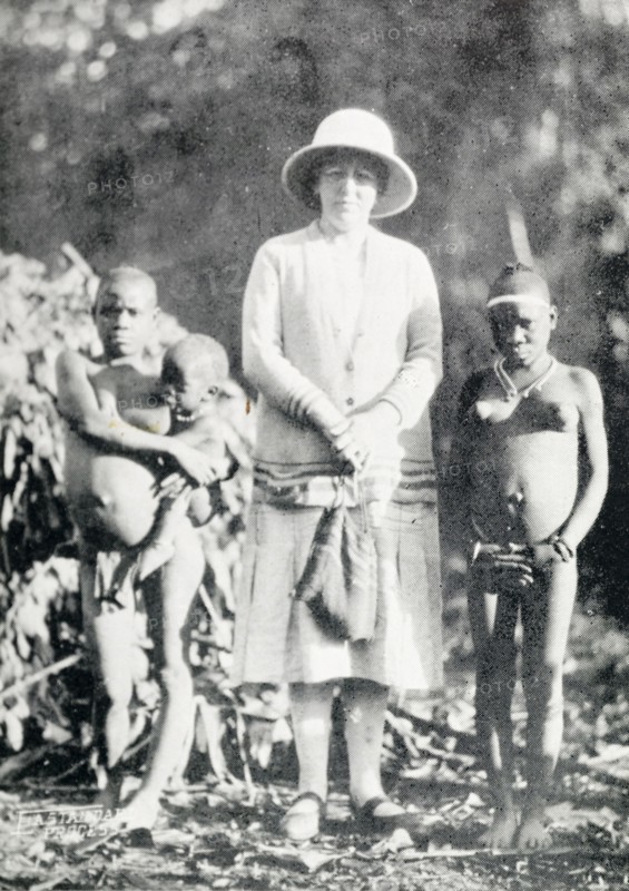 A British woman poses with "pigmy women". A British woman poses for a portrait in between two semi-naked African woman, who are identified in an original caption as "full grown pigmy (sic) women". British East Africa (Kenya), circa 1912. Aba, Abd Allah, Kenya, Eastern Africa, Africa.
