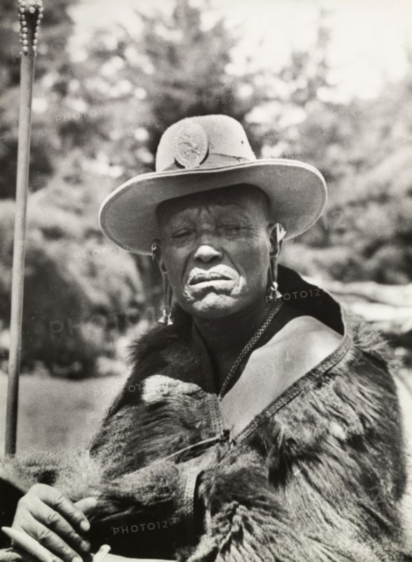 Chief Murigo. Portrait of Chief Murigo wearing traditional Kikuyu jewellery and holding a staff. He wears a Western-style, wide-brimmed hat with a badge depicting a lion, and an animal skin wrapped around his shoulders. Near Kakamega, Kenya, 1936. Kakamega, West (Kenya), Kenya, Eastern Africa, Africa.