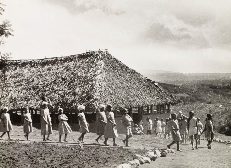 Kikuyu schoolchildren. A group of Kikuyu schoolchildren walk in single file towards a thatched roof school building in Nyeri. An original caption comments: "The modern world starts to break the mould of Kikuyu society, with Western (literary) education". Nyeri, Kenya, 1936. Nyeri, Central (Kenya), Kenya, Eastern Africa, Africa.