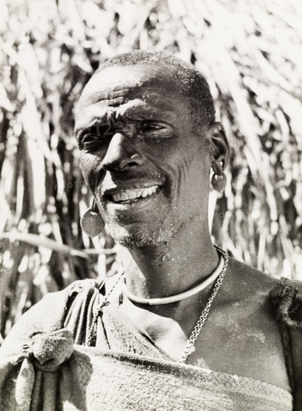 A Kikuyu witch doctor. Portrait of a smiling Kikuyu man, labelled in an original caption as a witch doctor. He wears a robe tied at the shoulder with earrings and a beaded necklace. South Nyeri, Kenya, 1936. Nyeri, Central (Kenya), Kenya, Eastern Africa, Africa.