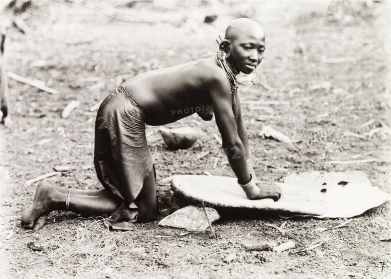 Grinding millet. A Kikuyu woman, naked from the waist up, kneels on the ground as she grinds millet on a flat board. Probably Nyeri, Kenya, circa 1936. Nyeri, Central (Kenya), Kenya, Eastern Africa, Africa.