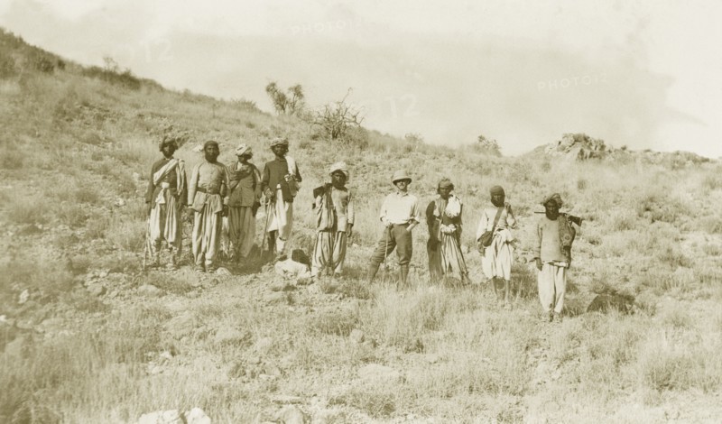 Walking sticks and a gun. Sir Henry Staveley Lawrence, Collector of Karachi, stands amongst a group of Indian men in a rocky landscape. Most of the men carry walking sticks and one of the group carries a gun slung over his shoulder. India (Pakistan), circa 1910. Pakistan, Southern Asia, Asia.