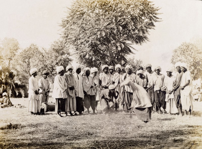 A wrestling match in Sukkur . Men in traditional Indian dress stand around a straw-covered patch of ground, watching as two men wrestle each other in a match. Sukkur, Sind, India (Sindh, Pakistan), circa 1908. Sukkur, Sindh, Pakistan, Southern Asia, Asia.