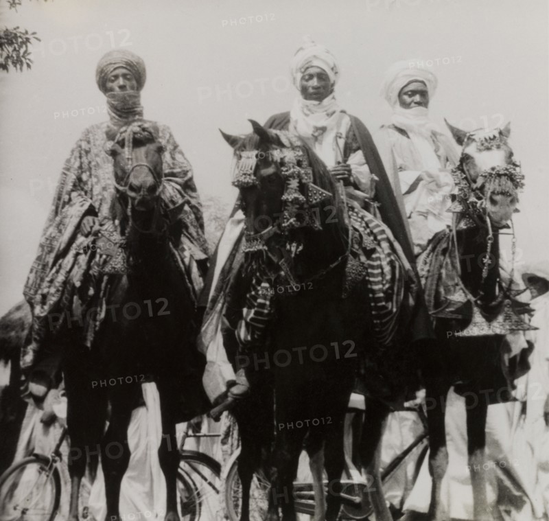 Nupe chiefs attend Sallah celebrations. Three Nupe chiefs dressed in ceremonial attire ride caparisoned horses during celebrations for Sallah, an Islamic event held to mark the end of Ramadan. Bida, Nigeria, October 1948. Bida, Niger, Nigeria, Western Africa, Africa.