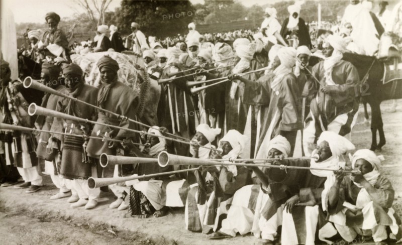 Nigerian trumpet players. A group of traditionally dressed Nigerian musicians assemble at an outdoor event to play a fanfare with long trumpets. Nigeria, circa 1955. Nigeria, Western Africa, Africa.