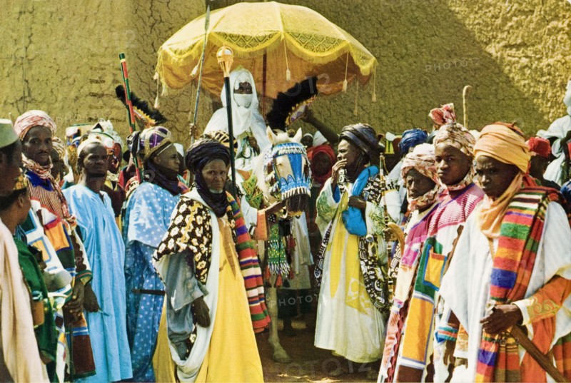 Alhaji Ado Bayero with his retinue. Portrait of Alhaji Ado Bayero (b.1930), the Emir of Kano, surrounded by members of his retinue who wear colourful ceremonial robes and head wraps. The Emir's face is covered and he sits beneath a ceremonial umbrella on the back of a caparisoned horse. Kano, Nigeria, circa 1975. Kano, Kano, Nigeria, Western Africa, Africa.