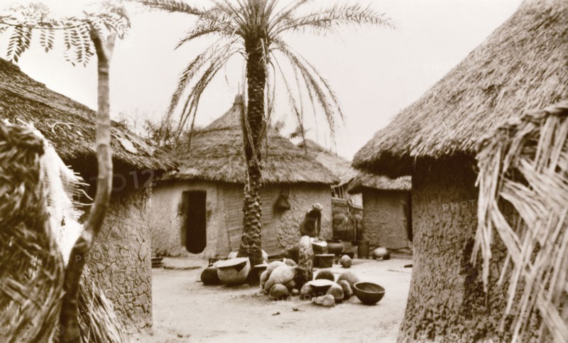 Interior of a Nigerian village. A woman undertakes domestic chores outdoors in the centre of a Nigerian village. The settlement comprises several round huts with mud walls and thatched roofs. Minna, Nigeria, circa 1940. Minna, Niger, Nigeria, Western Africa, Africa.