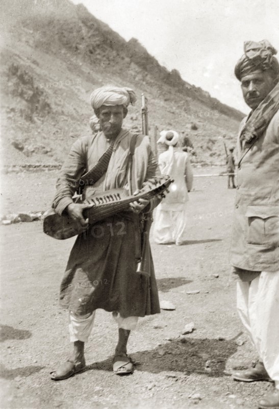 Playing a sarod at the border. An Indian man, armed with a rifle and wearing a bandolier, plucks a sarod (Indian stringed instrument) at a military checkpoint on the Afghanistan-India (Pakistan) border. India (Pakistan), circa 1930. Pakistan, Southern Asia, Asia.
