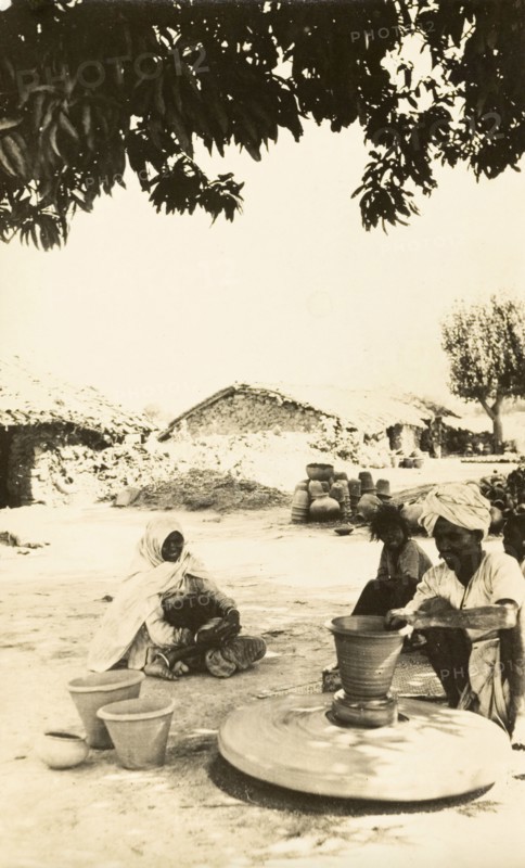 Indian craftsman at a potter's wheel. A craftsman sits beneath a shady tree as he sculpts a clay pot on a revolving wheel. India, circa 1920. India, Southern Asia, Asia.