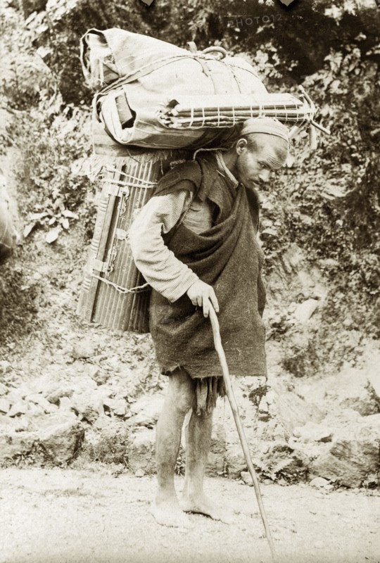 Burdened with luggage. An Indian man carries cumbersome bundles of luggage on his back as he walks barefoot along a road with the aid of a walking stick. India, circa 1920. India, Southern Asia, Asia.