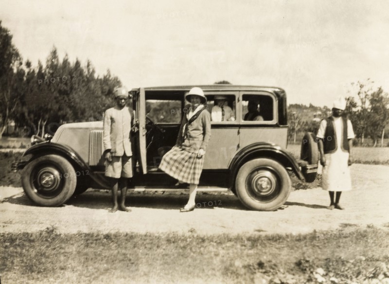 A head servant holds open a car door. An African head servant holds open the door of a car as a European woman prepares to climb into the front seat. Probably Nyasaland (Malawi), circa 1940. Malawi, Southern Africa, Africa.