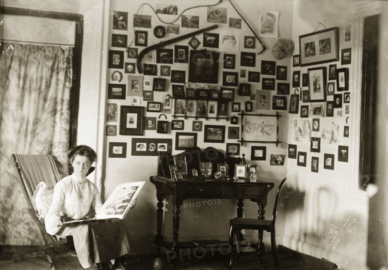 In the drawing room at Penshew House. A European woman browses through a photograph album in the drawing room of Penshew House. Behind her, the walls are covered with framed pictures and items of memorabilia. Bangalore, India, circa 1900. Bangalore, Karnataka, India, Southern Asia, Asia.