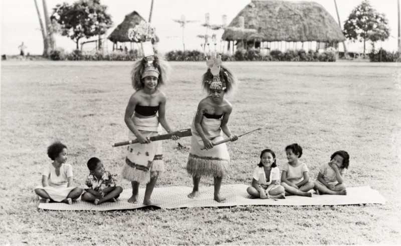 Samoan women perform a 'siva'. Two young Samoan women perform a 'siva' (dance) on a woven mat, watched by five young children. They are dressed in ceremonial attire comprising elaborate headdresses and fringed skirts. An original caption draws attention to a traditional Samoan 'fale' (house) in the distance, a thatched structure supported by upright beams. Western Samoa (Samoa), circa 1956. Samoa, Pacific Ocean, Oceania.