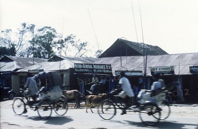 Cycle rickshaw drivers in Dibrugarh. Cycle rickshaw drivers whizz past shops with tin roofs on a commercial street in Dibrugarh. Dibrugarh, Assam, India, circa 1970. Dibrugarh, Assam, India, Southern Asia, Asia.
