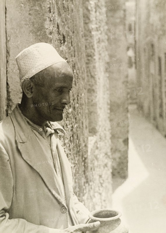 Profile shot of a Muslim man. Profile shot of a Muslim man wearing an embroidered kufi (cap). He leans against a stone wall on a narrow street, holding up a clay pot. Probably Lamu, Kenya, circa 1947. Lamu, Coast, Kenya, Eastern Africa, Africa.