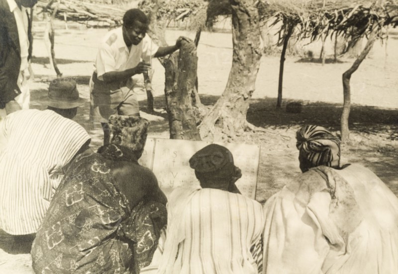 Adult literacy campaign, Gold Coast. A student teacher helps men to read from a board during a literacy class: part of a mass literacy campaign launched by the British government in 1951 to improve literacy skills amongst African adults, both in their own languages and in English. Northern Territories, Gold Coast (Northern Ghana), circa 1951., North (Ghana), Ghana, Western Africa, Africa.