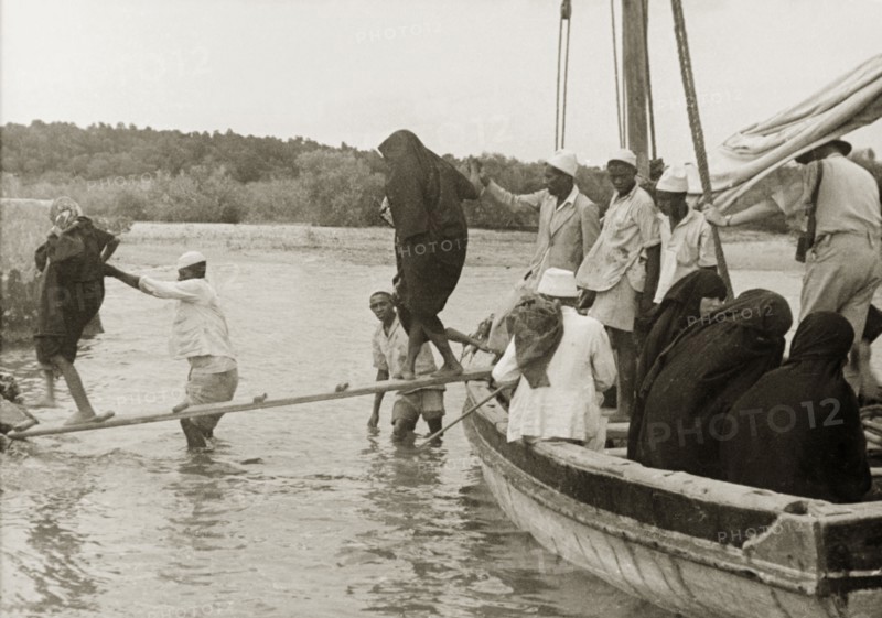 Disembarking at Mombasa Island. Arabic men assist a group of veiled Muslim women to cross a gangplank as they disembark from a sailing boat at Mombasa Island. Mombasa, Kenya, circa 1935. Mombasa, Coast, Kenya, Eastern Africa, Africa.