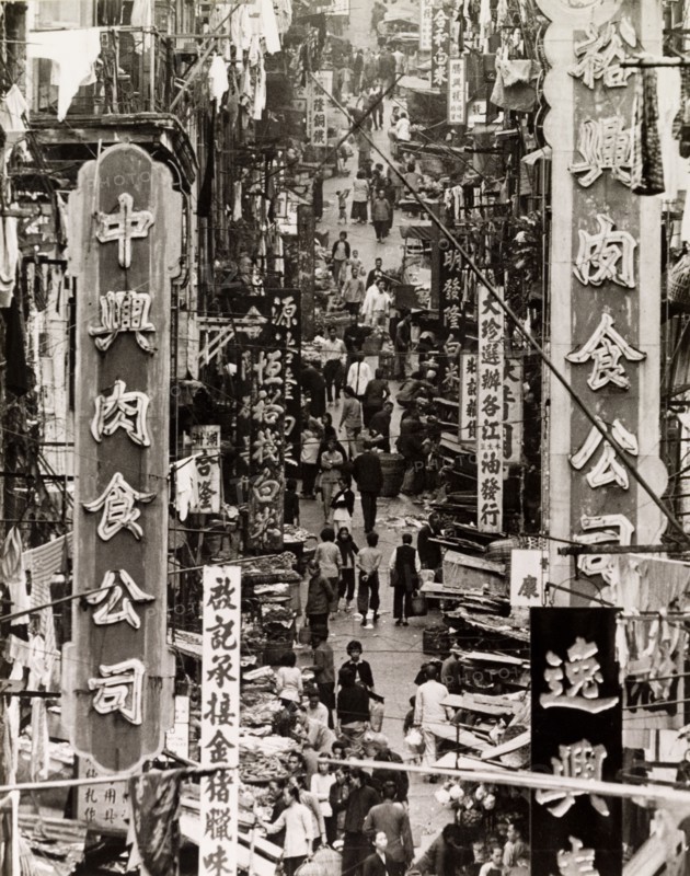 A busy side street in Hong Kong. Street traders vie for business on a city side street beneath a profusion of signs and advertisements written in Chinese script. Hong Kong, China, 1963. Hong Kong, Hong Kong, China, People's Republic of, Eastern Asia, Asia.