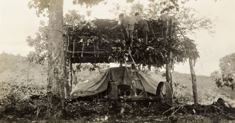Building a garage, Tanganyika. Three African men construct a thatched roof for a garage, under which sits a piece of agricultural machinery or a vehicle covered in tarpaulin. Moshi, Tanganyika Territory (Tanzania), circa 1930. Moshi, Kilimanjaro, Tanzania, Eastern Africa, Africa.