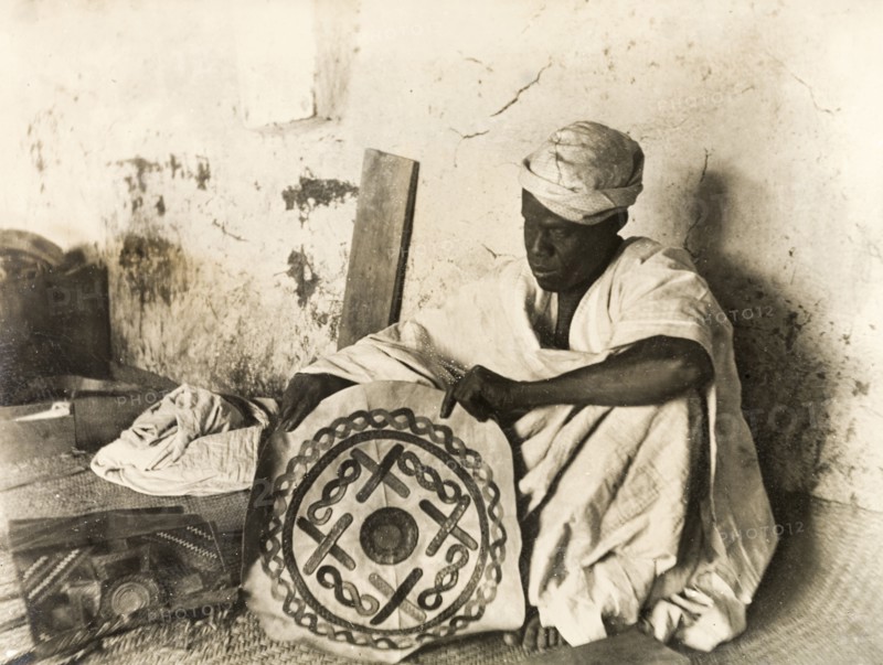 A Nigerian leatherworker. A Nigerian leatherworker sits in his workshop, holding up a decorative piece of leather that he has crafted. Probably Kano, Nigeria, circa 1915. Nigeria, Western Africa, Africa.