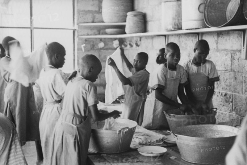 Laundry at a mission school. Kikuyu boys dressed in Western-style uniforms and aprons wash clothes in metal tubs inside a stone-walled laundry room. They are probably students of a Christian mission school, possibly the Church of Scotland mission school in south Nyeri. Kenya, circa 1936. Kenya, Eastern Africa, Africa.