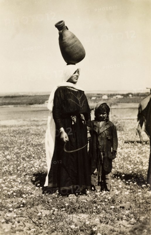 Bedouin woman carrying water pot. A bedouin woman stands with a young child in the Palestinian countryside, balancing a large clay pot on her head at a jaunty angle. British Mandate of Palestine (Middle East), circa 1942., Middle East, Asia.