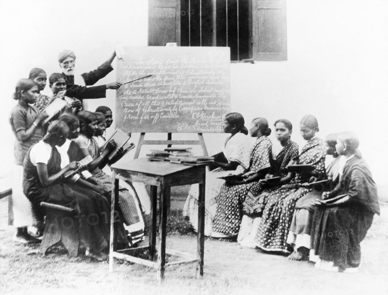 English lesson at a mission school, India. An Indian teacher points to a blackboard as he gives an English lesson outdoors at a school run by the London Missionary Society. He is surrounded by a class of Indian students who conscientiously copy text from the blackboard onto their slates. India, circa 1930. India, Southern Asia, Asia.