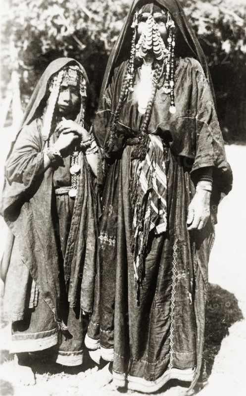 Portrait of two female 'bedouins'. Full-length portrait of an Arab woman and girl, identified in an original caption as ?bedouin(s)?. They wear long embroidered 'thobs', typical of Palestinian dress, with headscarves trailing down their backs and elaborate coin jewellery around their faces. British Mandate of Palestine (Middle East), circa 1938., Middle East, Asia.