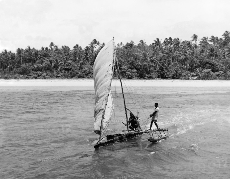 Manning a Fijian 'drua'. A 'drua' (Fijian catamaran) scuds across the waves away from a beach flanked by palm forest. Fiji, 1965. Fiji, Pacific Ocean, Oceania.
