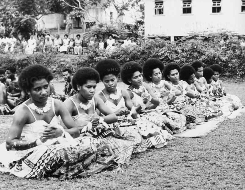 Women perform a 'vakamalolo'. Ten young Fijian women sit cross-legged in a line and clasp their hands together as they perform a 'vakamalolo' or sitting dance outdoors. All wear ceremonial costume including patterned 'sulus' (wraparound skirts) and typical Fijian hairstyles. Fiji, 1965. Fiji, Pacific Ocean, Oceania.