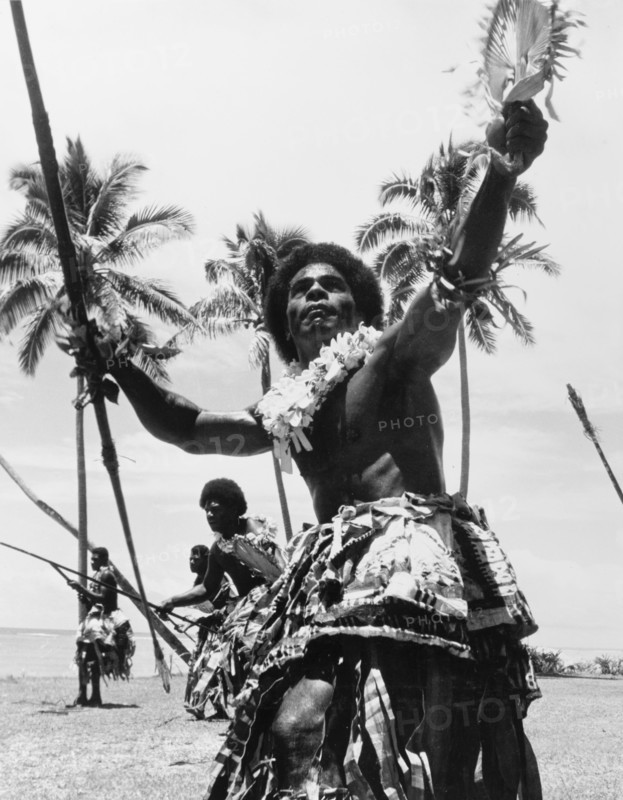 Men perform a 'meke wesi'. A group of men perform the 'meke wesi', a male war dance from Fiji that incorporates props such as spears and fans. Naked from the waist up, they wear ceremonial costume including layered 'sulus' (wraparound skirts) and flowered leis. Fiji. 1965. Fiji, Pacific Ocean, Oceania.