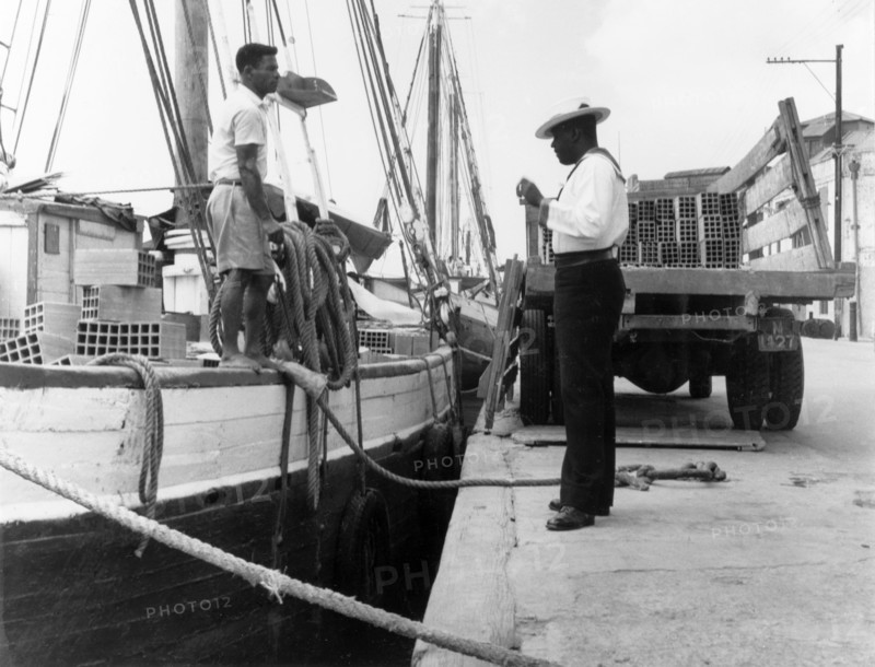 Policing the Careenage. A harbour police officer talks to a man unloading bricks from a sail boat onto a truck as he patrols the Careenage. An original caption comments: "Harbour policemen in the (sailor's) uniform of... Nelson's day are always a target for photographers". Bridgetown, Barbados, 1965. Bridgetown, St Michael, Barbados, Caribbean, North America .