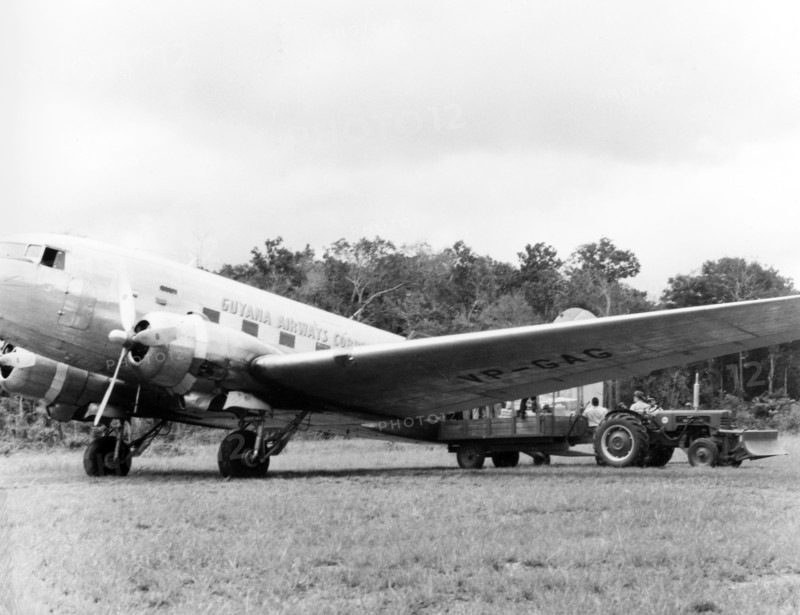 A Guyana Airways Corporation aeroplane . A Guyana Airways Corporation aeroplane sits in a grassy field as it is loaded with cargo from the back of a tractor. An original caption comments that the aircraft was "en route to the interior". British Guiana (Guyana), 1965., Guyana, South America, South America .