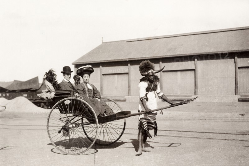 Zulu rickshaw driver, Durban. A Zulu rickshaw driver takes a European couple for a ride. He is dressed in a traditional costume with a flamboyantly horned and feathered headdress. Durban, South Africa, 1904. Durban, KwaZulu Natal, South Africa, Southern Africa, Africa.