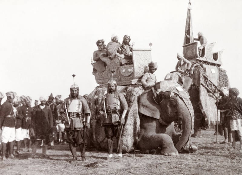 Bhopal State procession at Coronation Durbar, 1903. Two armoured soldiers of Bhopal State stand beside a kneeling caparisoned elephant, on whose back three Indian dignitaries are sitting in an ornate howdah. A second elephant approaches the group, ready to pick up its passengers and join a procession at Edward VII's Coronation Durbar. Delhi, India, circa 1 January 1903. Delhi, Delhi, India, Southern Asia, Asia.