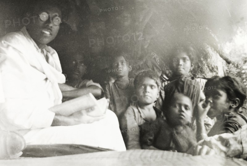 Reading to young children, India. A Methodist evangelist reads aloud from a book to a group of young children, possibly the children of coffee plantation workers, who are gathered next to him under the eaves of a thatched hut. Pandaravalli, Mysore State (Karnataka), India, 1937., Karnataka, India, Southern Asia, Asia.