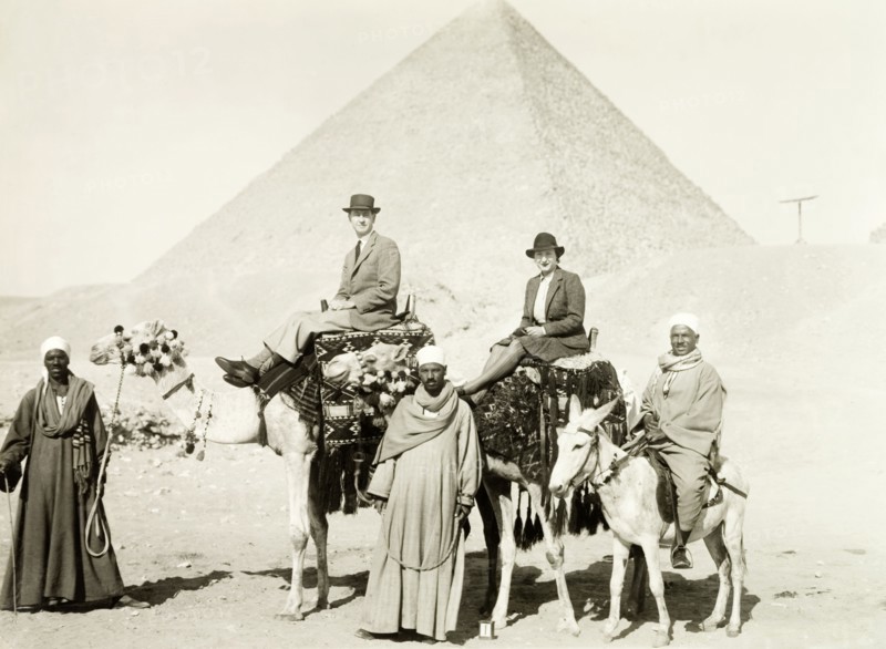 Riding camels at the Great Pyramid. William Ryder McGeagh, District Commissioner in Jerusalem, and his wife ride a pair of caparisoned camels in front of the Great Pyramid. They are guided by three Egyptian men in long robes: one of whom rides a donkey. Giza, Egypt, January 1939. Giza, Giza, Egypt, Northern Africa, Africa.