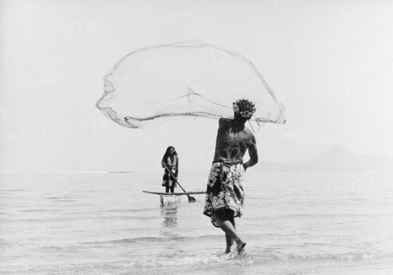 Fishing with a cast net. A Tahitian man wearing a printed 'pareu' (sarong) hurls a cast net into shallow waters off the coast. Opposite him, a woman wearing a flowered lei pilots a canoe equipped with an outrigger. Tahiti, French Polynesia, 1965., Windward Islands (including Tahiti), French Polynesia, Pacific Ocean, Oceania.