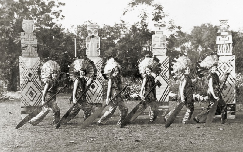 Chorus line act at a garden party. Members of the cast of 'Flying Fragments' performed by the Moradabad Amateur Dramatic Society at the Railway Institute. Several women perform a chorus line act at a garden party, dressed in traditional Native American costume. They strike a pose in front of four artificial totem poles, each holding a large oar with both hands as part of the Red Indian Canoe Dance. Moradabad, United Provinces (Uttar Pradesh), India, 12 February 1934. Moradabad, Uttar Pradesh, India, Southern Asia, Asia.