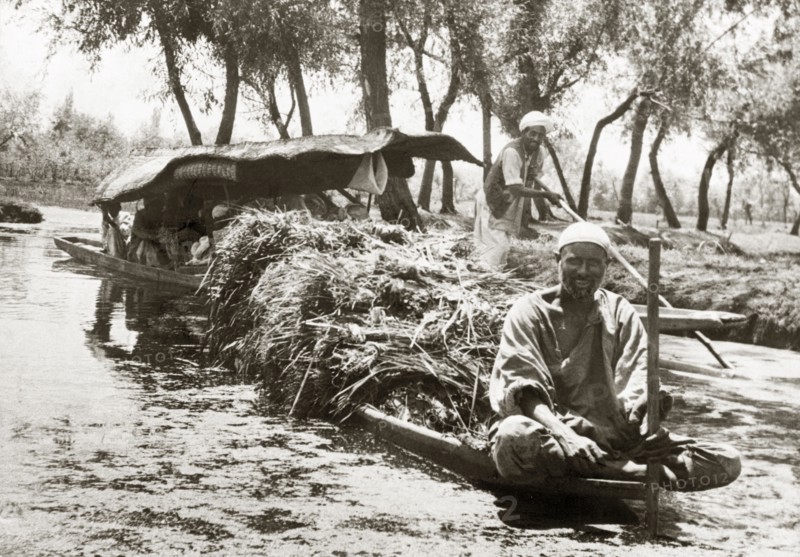 A canal dredger in Srinagar. A canal dredger sits cross-legged on the bow of a long canoe, which is piled high with cut reeds. Srinagar, Jammu and Kashmir, India, 1934. Srinagar, Jammu and Kashmir, India, Southern Asia, Asia.
