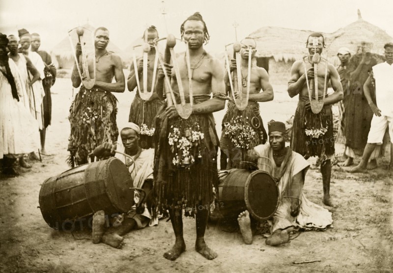 Nigerian musicians and hoe dancers. Five Nigerian hoe dancers, dressed in grass skirts with bells and bangles, pose for a group portrait holding their hoes. Two musicians sit on the floor amongst the group, playing the 'ganga' (drums) with curved sticks. Nigeria, circa 1915. Nigeria, Western Africa, Africa.