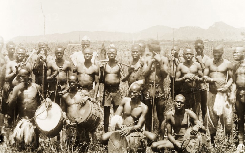 Labourers and musicians, Nigeria. A group of labourers assemble for a group portrait around four musicians who hold 'gangas' (drums) with curved sticks. Bauchi State, Nigeria, circa 1915., Bauchi, Nigeria, Western Africa, Africa.