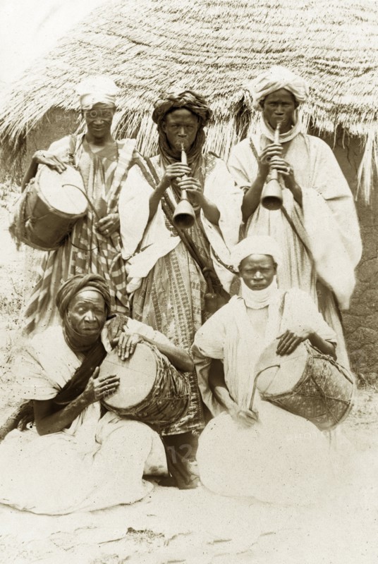 Hausa musical band. Five Hausa musicians pose for a group portrait with their instruments. Two men play the 'algaita' (oboe), whilst the other three beat 'gangas' (drums) using curved sticks. Nigeria, circa 1915. Nigeria, Western Africa, Africa.