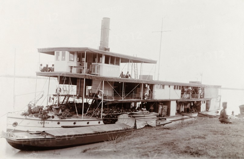 A paddle steamer named 'Sarota', Nigeria. A paddle steamer named 'Sarota' is moored to a riverbank in Nigeria. Several passengers are visible onboard, waiting for the vessel to leave shore. Nigeria, circa 1910. Nigeria, Western Africa, Africa.